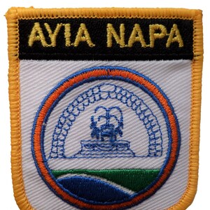 May include: Embroidered patch with the text "AYIA NAPA" in gold letters on a black background. The patch features a circular design with a blue and orange border, depicting a fountain and archway. The patch has a yellow border.
