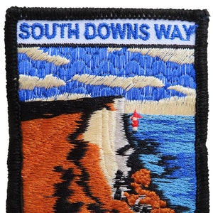 May include: Embroidered patch with a black border and the text "SOUTH DOWNS WAY" in blue. The patch features a scenic image of a cliff overlooking a blue body of water with a red buoy.