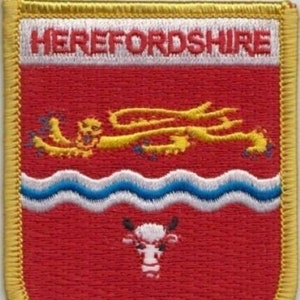 May include: An embroidered patch featuring the coat of arms of Herefordshire. The patch is red with a white border and features a yellow dragon, blue waves, and a white bull. The text "Herefordshire" is embroidered in white on a white background.