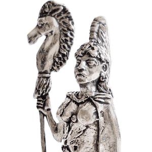 Op de afbeelding: Een zilveren beeldje van een vrouw die een staf vasthoudt met een paardenkop erop. De vrouw draagt een mantel en een rok. De basis van het beeldje is versierd met een zon en een Keltisch knoop.