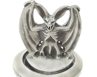 Puede incluir: Un tapón de botella de peltre con un diseño de murciélago detallado con las alas extendidas. El murciélago tiene una expresión amenazante con dientes y garras afiladas. El tapón está unido a una base de corcho.