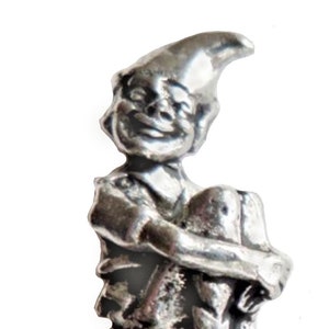 Pode incluir: Uma figura de estanho prateado de um gnomo sentado em um cogumelo. O gnomo está usando um chapéu pontudo e tem um rosto sorridente.