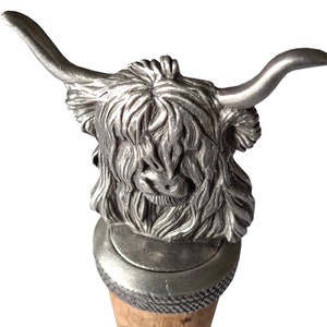 Puede incluir: Un tapón de botella de plata con forma de vaca Highland con base de corcho. La cabeza de la vaca está mirando hacia adelante con los cuernos apuntando hacia afuera.