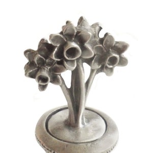 以下が含まれることがあります： 3つの水仙の花のデザインが施された銀製のボトルストッパー。ストッパーはピューター製で、コルクのベースが付いています。