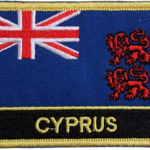 Könnte beinhalten: Ein blauer gestickter Aufnäher mit der Flagge von Zypern. Die Flagge zeigt im oberen linken Eck eine Union Jack und rechts ein rotes Löwenemblem. Der Aufnäher hat einen schwarzen Rand mit goldenen Nähten und das Wort "CYPRUS" in goldenen Buchstaben.