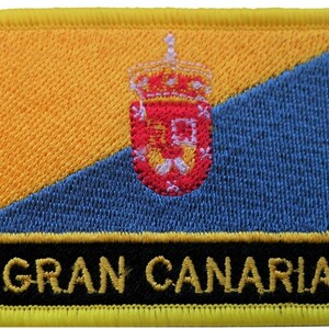 Könnte beinhalten: Gestickter Aufnäher mit der Flagge von Gran Canaria. Das Design umfasst eine gelbe und blaue diagonale Teilung, ein Wappen und den Text "GRAN CANARIA" in Gold auf einem schwarzen Balken. Der Aufnäher hat einen gelben Rand.