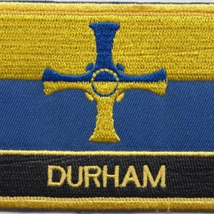 May include: An embroidered patch featuring a yellow and blue design with a gold cross and the word "DURHAM" in gold lettering on a black background.
