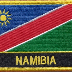 Pode incluir: Um remendo bordado com a bandeira da Namíbia, com um design amarelo, vermelho, verde e azul. O remendo tem um sol branco no canto superior esquerdo e a palavra "NAMIBIA" em letras amarelas sobre um fundo preto.