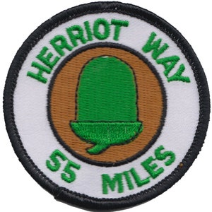 Può includere: Toppa rotonda bianca ricamata con cuciture nere. La toppa presenta un cerchio marrone con una ghianda verde al centro. Il testo "Herriot Way 55 Miles" è ricamato in verde attorno al cerchio marrone.