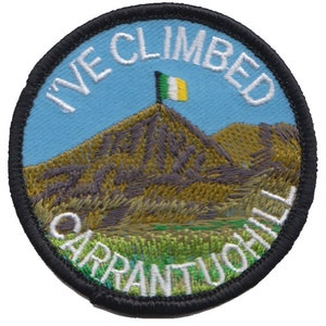 May include: A round, embroidered patch with a black border. The patch features a blue sky with a green, white, and orange flag on top of a brown mountain. The text "I've Climbed Carrantuohill" is embroidered in white around the edge of the patch.