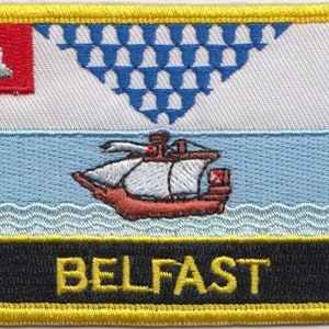 Op de afbeelding: Een geel geborduurd patch met het wapen van de stad Belfast. Het wapen toont een rode bel, een blauw-wit geruit patroon, een schip dat op blauw water vaart en het woord "BELFAST" in gele letters.