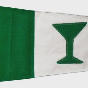 May include: A green and white pennant flag with a green martini glass design. The flag has a green section on the left, a white center with the martini glass, and a green section on the right. It has grommets and ties.