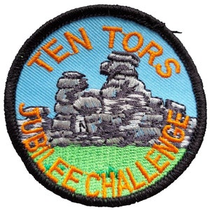May include: A round, blue and green embroidered patch with the text "Ten Tors Jubilee Challenge" in orange lettering around a gray embroidered image of a rock formation.