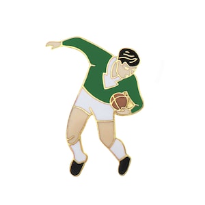 May include: A gold-toned enamel pin of a person in a green jersey and white shorts, holding a brown ball, in a running pose.