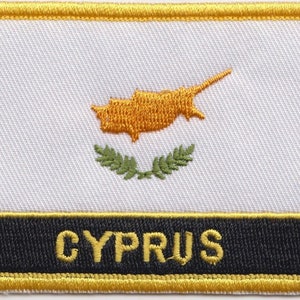May include: A white embroidered patch with a yellow outline. The patch features a yellow map of Cyprus with a green olive branch wreath below. The text "CYPRUS" is embroidered in yellow on a black background at the bottom of the patch.
