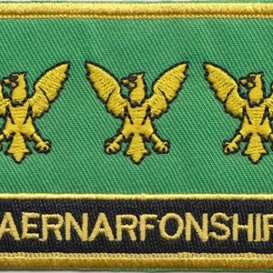 Könnte beinhalten: Ein gestickter Aufnäher in Grün und Gold mit drei gelben Adlern und dem Text "CAERNARFONSHIRE" in Schwarz und Gold.