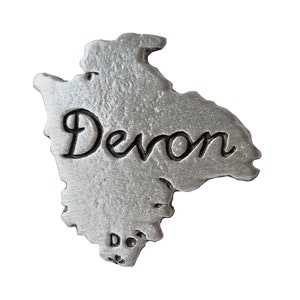 Könnte beinhalten: Eine silberne Zinn-Umrandung des englischen Countys Devon. Das Wort "Devon" ist in die Umrandung eingraviert.