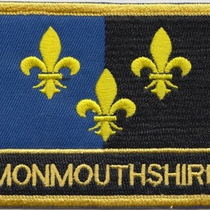 Könnte beinhalten: Gestickter Aufnäher mit dem Wort "MONMOUTHSHIRE" in Gold. Der Aufnäher ist in blaue und schwarze Hälften unterteilt, mit drei gelben Fleur-de-Lis-Symbolen. Der Rand ist goldfarben.