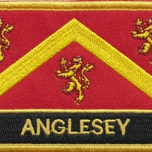 May include: Embroidered patch featuring a red background with a yellow chevron and three yellow lions. The word "ANGLESEY" is embroidered in yellow on a black banner at the bottom. The patch has a yellow border.
