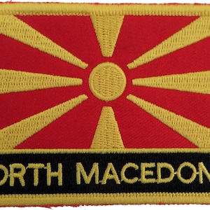 May include: Embroidered patch featuring the flag of North Macedonia. The flag is red with a yellow sun in the center and eight yellow rays extending from the sun. The text "NORTH MACEDONIA" is written in yellow letters on a black background below the flag.