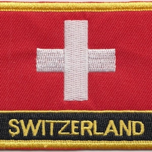 Pode incluir: Um remendo bordado vermelho com uma cruz branca e a palavra "SUÍÇA" em letras amarelas sobre um fundo preto. O remendo tem uma borda dourada.