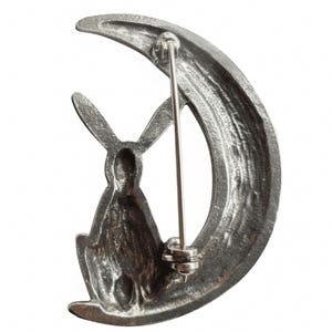 May include: A silver-toned brooch featuring a rabbit sitting on a crescent moon. The brooch has a pin back.