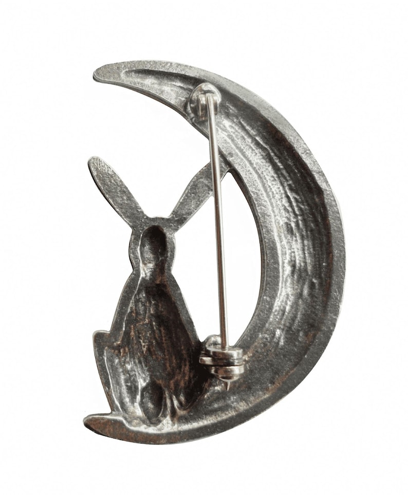 May include: A silver-toned brooch featuring a rabbit sitting on a crescent moon. The brooch has a pin back.