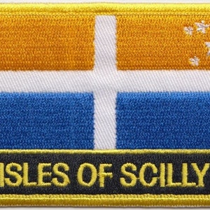 Könnte beinhalten: Ein gestickter Aufnäher in Gelb, Weiß und Blau mit dem Text "ISLES OF SCILLY" in gelben Buchstaben auf schwarzem Hintergrund. Der Aufnäher zeigt ein weißes Kreuz auf gelbem Hintergrund mit fünf weißen Sternen in der oberen rechten Ecke.