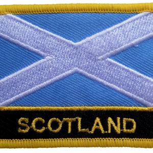 May include: A blue and white embroidered patch with a white cross on a blue background. The patch has a black border with gold trim and the word "SCOTLAND" in gold lettering.