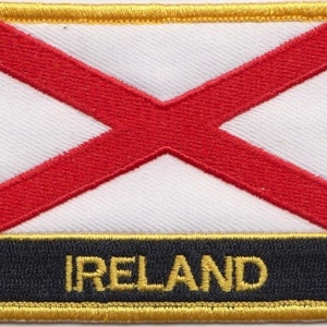 Könnte beinhalten: Ein weißer Patch mit einem roten diagonalen Kreuz, umgeben von einem goldenen Rand. Der Text "IRELAND" ist in goldenen Buchstaben auf schwarzem Hintergrund unter dem Patch geschrieben.
