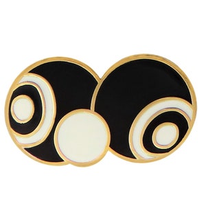 Puede incluir: Un broche de tono dorado con dos círculos negros y blancos. Los círculos tienen anillos concéntricos de blanco y negro, con un círculo blanco más pequeño en el centro de cada uno.