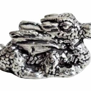 Puede incluir: Una figurita de peltre de color plateado de un pequeño dragón enroscado en posición de dormir. El dragón tiene escamas y alas detalladas.