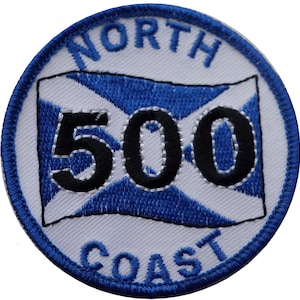 Könnte beinhalten: Ein runder weißer gestickter Aufnäher mit blauem Rand. Der Aufnäher zeigt eine blau-weiße schottische Flagge mit der Zahl 500 in schwarzer Schrift. Die Worte "NORTH COAST" sind in blauer Schrift um die Flagge herum gestickt.