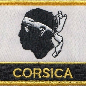 May include: A white patch with a black silhouette of a head wearing a white headband and a white sash. The patch has a black border with the word "CORSICA" in yellow lettering.