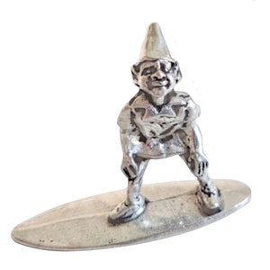 Puede incluir: Una figurita de plata de un gnomo con un sombrero puntiagudo, de pie sobre una tabla de surf. El gnomo tiene una expresión traviesa en su rostro.