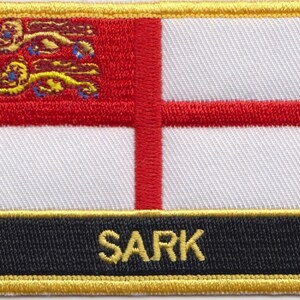 Op de afbeelding: Een geborduurde patch in rood, wit en goud met het woord "SARK" in gouden letters op een zwarte achtergrond. De patch heeft een rood kruis op een witte achtergrond met een rood en gouden leeuwenembleem in de linkerbovenhoek.