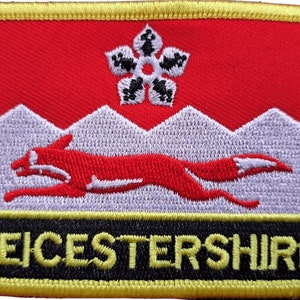 Pode incluir: Um remendo bordado vermelho, branco e preto com uma raposa branca correndo sobre uma cordilheira branca. Uma estrela negra de seis pontas está acima das montanhas. O texto "LEICESTERSHIRE" está em letras pretas sobre um fundo preto na parte inferior do remendo.