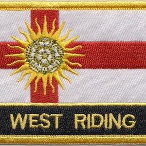 Könnte beinhalten: Ein weißer Patch mit einem roten Kreuz und einer gelben Sonne mit weißem Zentrum. Der Patch hat einen schwarzen Rand mit dem Text "WEST RIDING" in gelben Buchstaben.