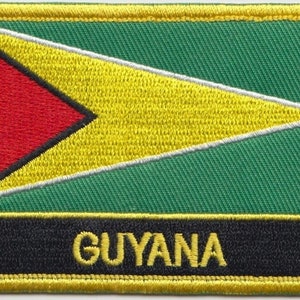 Op de afbeelding: Een geborduurde patch met de vlag van Guyana, met een groene achtergrond, een gele driehoek met een rode driehoek erin en het woord "GUYANA" in gele letters op een zwarte achtergrond.