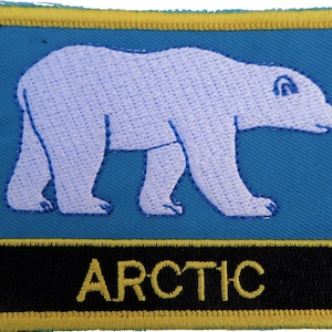 May include: An embroidered patch featuring a white polar bear walking across a blue background. The text "ARCTIC" is written in yellow letters on a black background at the bottom of the patch.