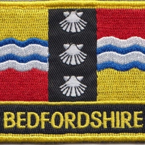 Op de afbeelding: Geborduurde patch met het ontwerp van de vlag van Bedfordshire. De patch heeft een gele rand en een zwarte streep met het woord "BEDFORDSHIRE". Het ontwerp bevat golvende lijnen en drie witte sint-jakobsschelpen.