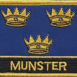 Może przedstawiać: Haftowana naszywka z trzema złotymi koronami na niebieskim tle, ze słowem "MUNSTER" złotymi literami na czarnym banerze. Naszywka ma złotą obwódkę.