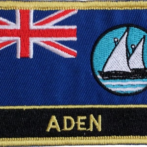 Könnte beinhalten: Ein blauer und weißer gestickter Aufnäher mit einer Union Jack in der oberen linken Ecke und einem weißen Segelboot in einem blauen Kreis in der oberen rechten Ecke. Der Aufnäher hat einen schwarzen Rand mit dem Wort "ADEN" in goldenen Buchstaben.