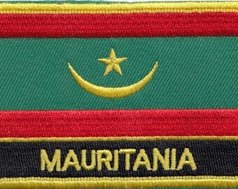Może przedstawiać: Naszywka haftowana w kolorze zielonym, czerwonym i czarnym z złotym półksiężycem i gwiazdą. Tekst "MAURITANIA" jest wyszyty złotem na czarnym tle.