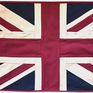 Pode incluir: Uma bandeira retangular de tecido com o design da Union Jack em tons de bordô profundo, azul-marinho e branco sujo. A bandeira inclui uma faixa de reforço de lona bege no lado esquerdo com dois ilhós metálicos para montagem e costuras.