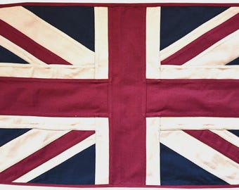 May include: A rectangular fabric flag featuring the Union Jack design in deep burgundy, navy blue, and off-white. The flag includes a reinforced beige canvas header on the left side with two metal grommets for mounting, showcasing stitched seams.