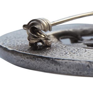 May include: Close-up of a silver-toned brooch with a detailed design. The brooch has a pin and catch closure.