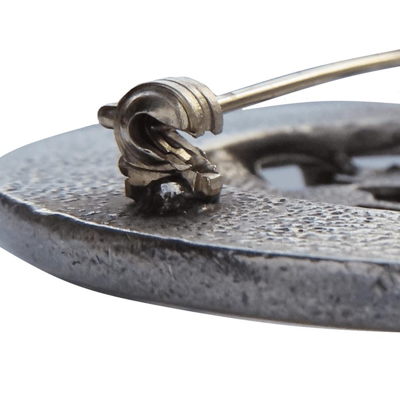 May include: Close-up of a silver-toned brooch with a detailed design. The brooch has a pin and catch closure.