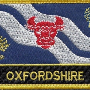 Könnte beinhalten: Ein blauer und weißer gestickter Aufnäher mit einem roten Stierkopf, einem gelben Baum und einer gelben und blauen Weizengarbe. Der Aufnäher hat einen schwarzen Hintergrund mit dem Wort "OXFORDSHIRE" in gelben Buchstaben.