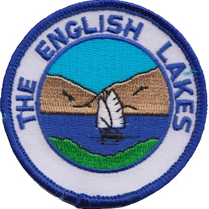 Puede incluir: Un parche bordado azul y blanco con el texto "The English Lakes" en un círculo. El parche presenta un velero en un lago con montañas en el fondo.